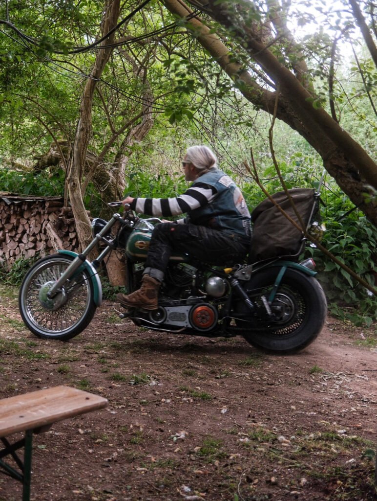 Eine ältere Person mit langen grauen Haaren, einem gestreiften Hemd und Stiefeln fährt auf einem alten Motorrad auf einem unbefestigten Weg, der von üppigem Grün und Bäumen umgeben ist. Im Hintergrund ist ein Stapel Brennholz zu sehen.