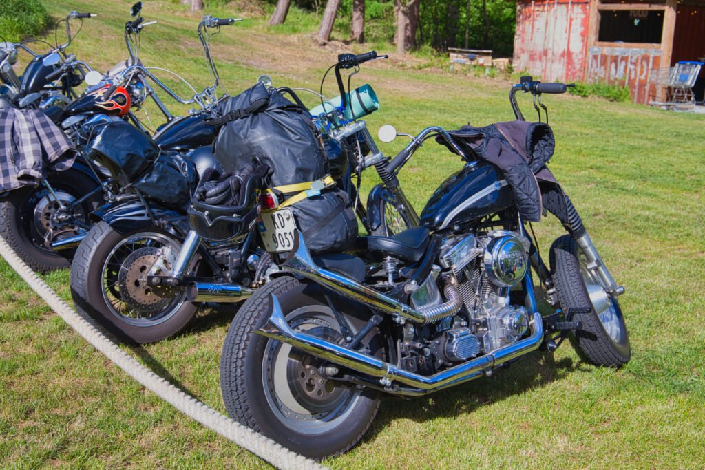 Drei geparkte Motorräder mit Taschen und Jacken liegen auf einer Wiese in der Nähe eines weißen Seils, mit Bäumen und einem rustikalen roten Gebäude im Hintergrund an einem sonnigen Tag.