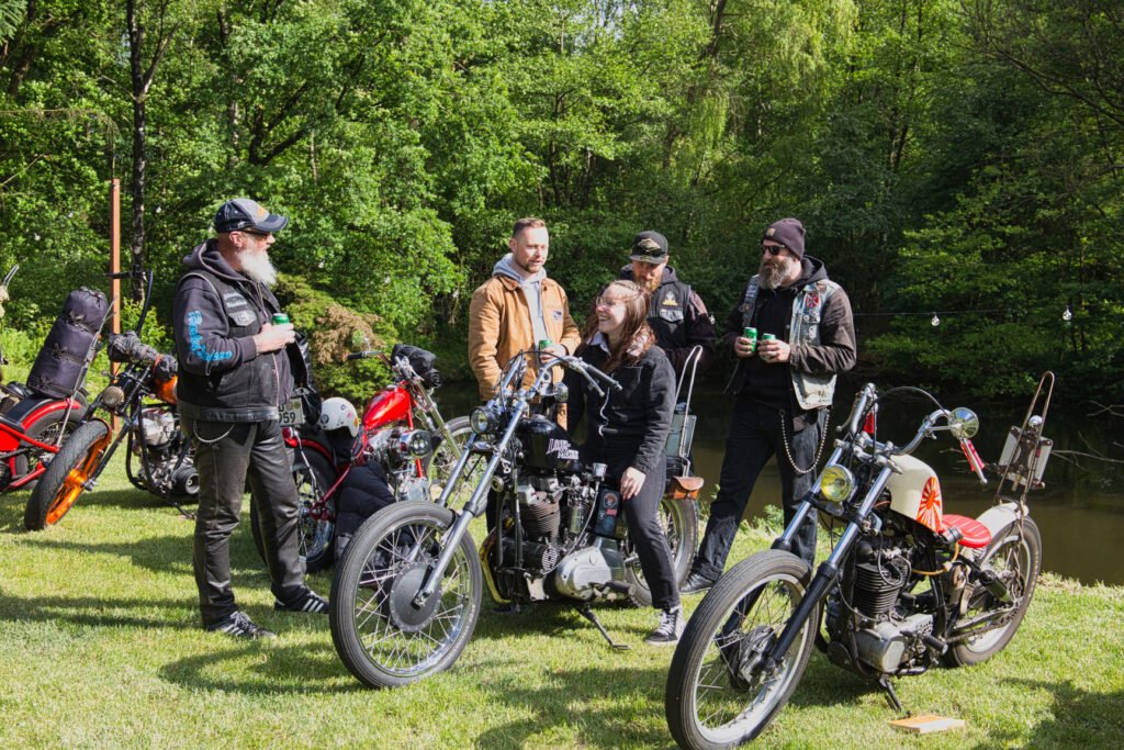 Eine Gruppe von sechs Personen, einige stehend, andere auf Motorrädern sitzend, plaudert und lacht auf einer Wiese mit Bäumen und geparkten Motorrädern im Hintergrund.