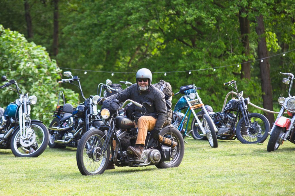 Eine Person mit Helm und Sonnenbrille fährt auf einem Oldtimer-Motorrad auf einer Wiese, umgeben von mehreren geparkten Motorrädern, mit grünen Bäumen im Hintergrund.