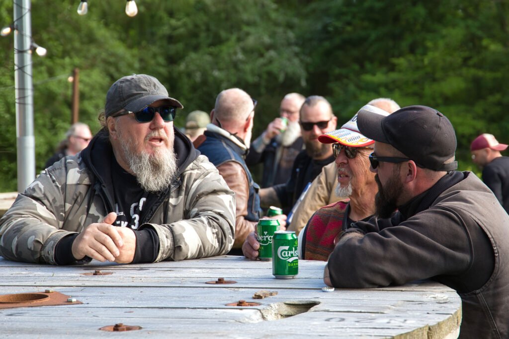 Drei Männer in legerer Outdoor-Kleidung und mit Sonnenbrille sitzen an einem runden Holztisch, unterhalten sich und halten Bierdosen in der Hand, während andere Menschen und Grünflächen im Hintergrund verschwimmen.