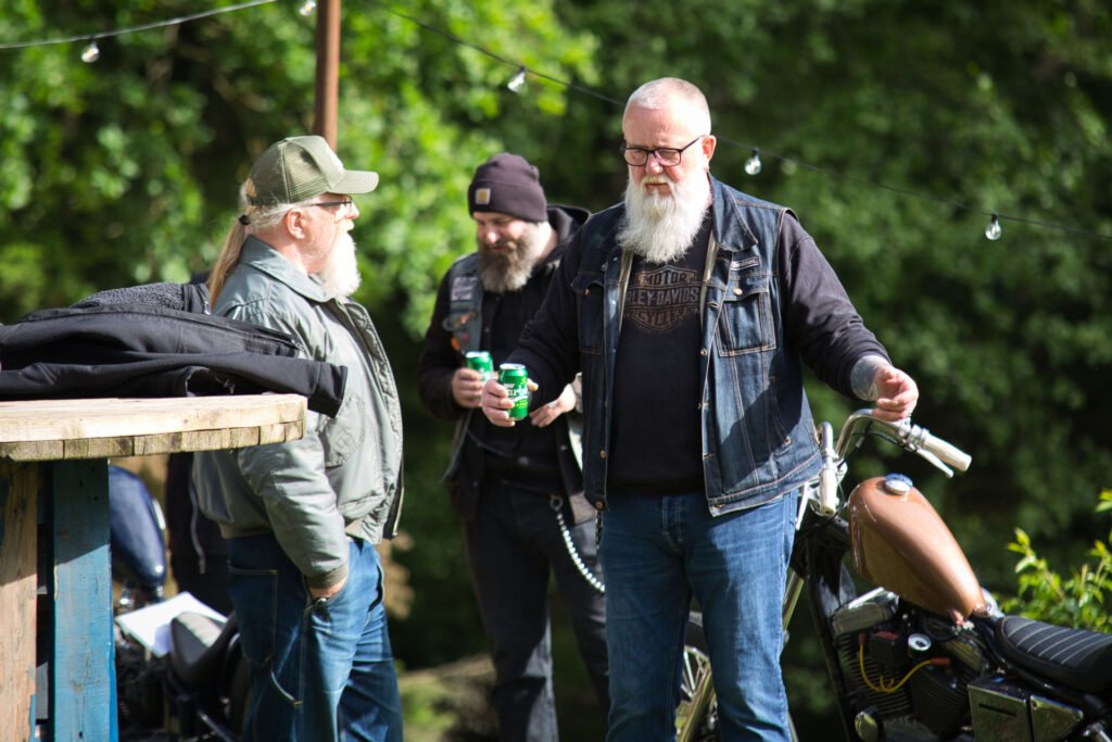 Drei Männer mit Bärten und legerer Kleidung stehen im Freien neben einem Motorrad, halten Getränke in der Hand und unterhalten sich. Im Hintergrund sind Bäume und Lichterketten zu sehen, die eine entspannte, gesellige Atmosphäre schaffen.