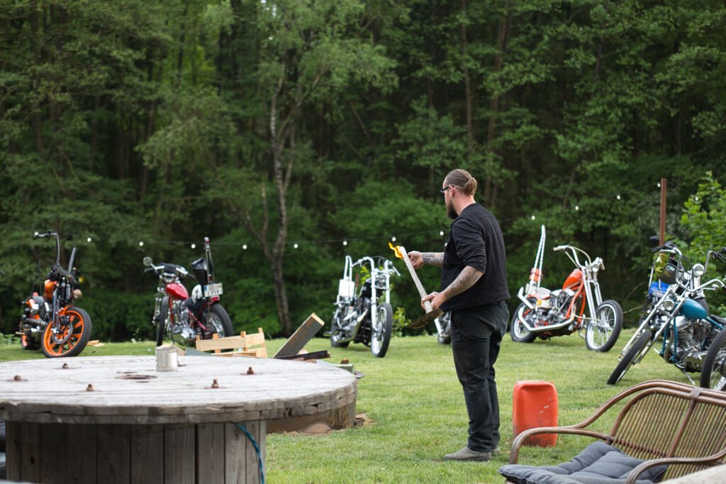 Ein Mann mit einer Axt steht auf einer Wiese in der Nähe einer Feuerstelle, im Hintergrund sind mehrere Motorräder geparkt und dahinter stehen Bäume. In der Nähe befinden sich eine große Holzspule und Gartenstühle.