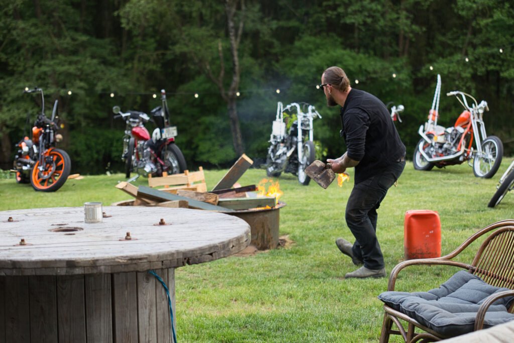 Ein Mann legt Holz für eine Feuerstelle auf einer Wiese nach. Im Hintergrund sind mehrere Custom-Motorräder geparkt, über denen Lichterketten hängen. Im Vordergrund stehen ein hölzerner Spulentisch und ein Korbsessel mit Kissen.