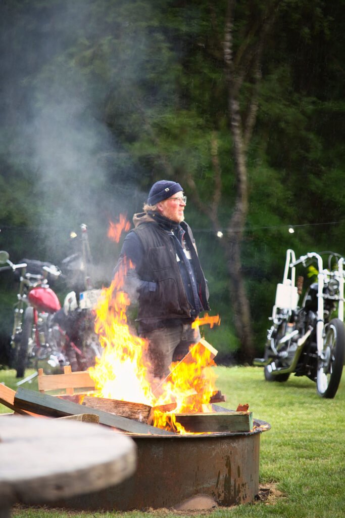 Eine Person in schwarzer Jacke und Mütze steht in der Nähe einer großen Feuerstelle mit hellen Flammen. Im Hintergrund sind Motorräder und Grünpflanzen zu sehen, und die Szene scheint im Freien zu spielen.