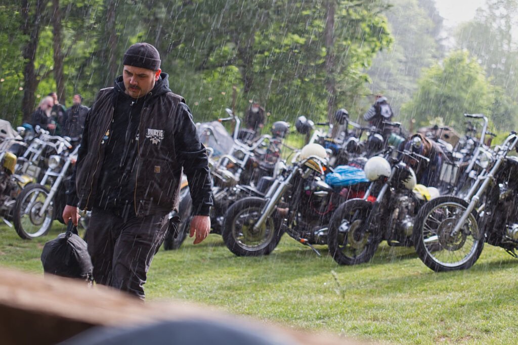Ein dunkel gekleideter Mann geht im Regen auf einer Wiese spazieren und trägt eine Tasche, im Hintergrund sind viele geparkte Motorräder und Bäume zu sehen.