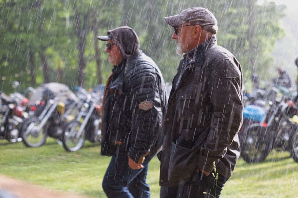 Zwei Männer in dunklen Jacken und Hüten gehen durch starken Regen bei einer Versammlung im Freien, während im Hintergrund mehrere Motorräder auf grünem Gras geparkt sind.