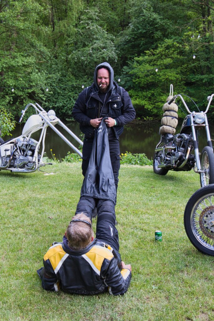 Eine Person in schwarzer Motorradkleidung steht im Gras und hält die Beine einer anderen liegenden Person, die eine schwarz-gelbe Jacke trägt. Im Hintergrund sind Motorräder und ein Teich zu sehen, die Szene ist von Bäumen umgeben.