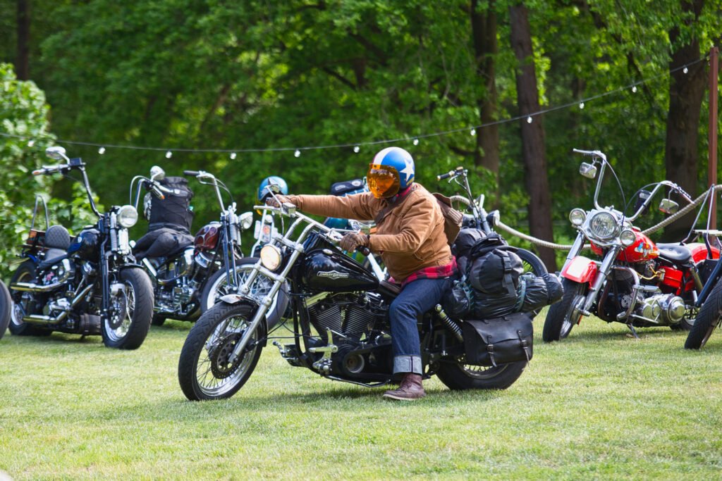Eine Person, die einen blau-orangen Helm und eine braune Jacke trägt, fährt auf einem schwarzen Motorrad auf einer Wiese, mit mehreren anderen geparkten Motorrädern und grünen Bäumen im Hintergrund.