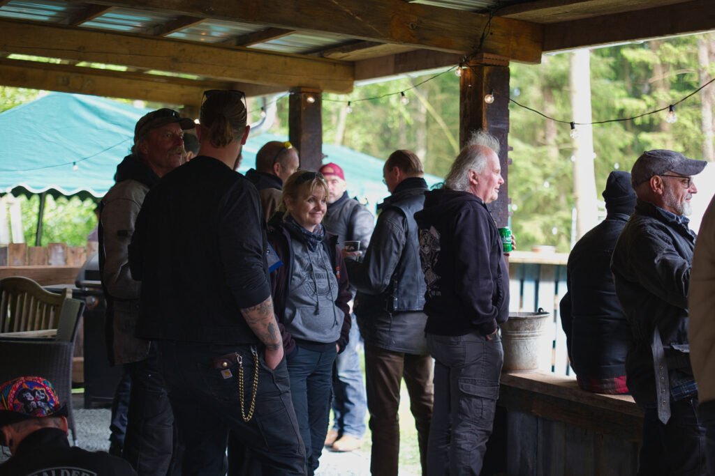 Eine Gruppe von Menschen, meist Männer, steht unter einem Holzunterstand mit Lichterketten und unterhält sich. Einige von ihnen tragen Jacken und Hüte. Im Hintergrund sind grüne Bäume und ein Zelt im Freien zu sehen.