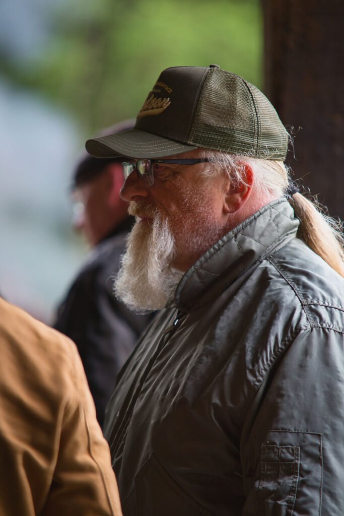 Ein bärtiger Mann mit Brille und Pferdeschwanz, der eine grüne Mütze und eine graue Jacke trägt, steht im Freien. Der Hintergrund ist leicht verschwommen und zeigt eine weitere Person und grünes Laub.