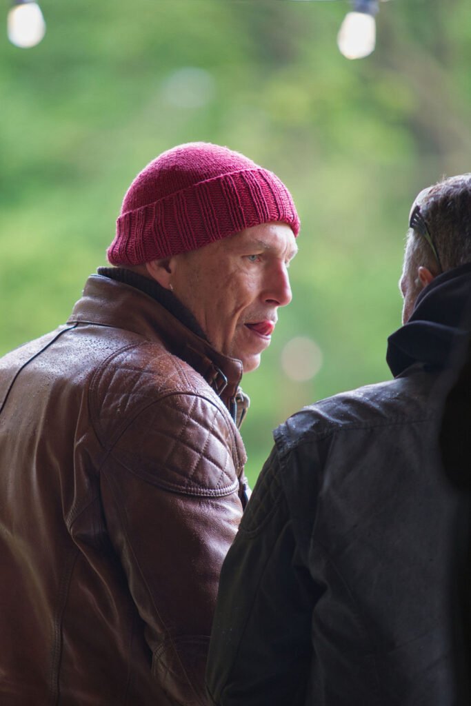 Eine Person mit roter Strickmütze und brauner Lederjacke unterhält sich mit einer anderen Person im Freien, wobei das Grün im Hintergrund verschwimmt.