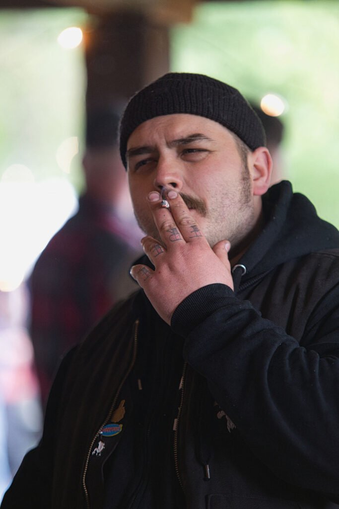 Ein Mann mit schwarzer Mütze und dunkler Jacke raucht eine Zigarette und hält sie mit tätowierten Fingern an seine Lippen. Er blickt nachdenklich nach vorne, hinter ihm ein verschwommener Hintergrund im Freien.