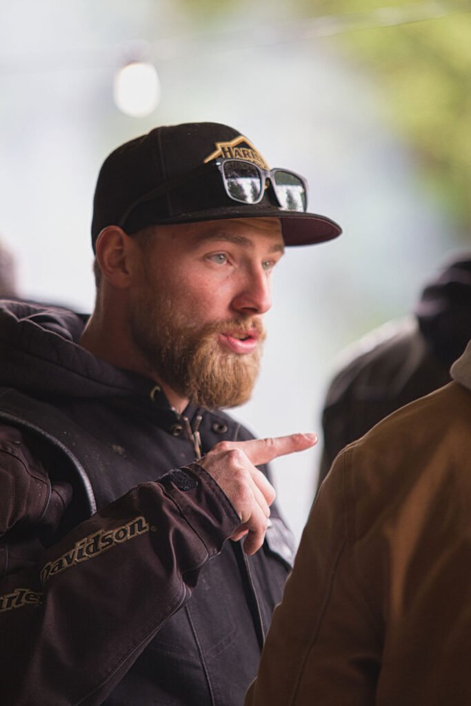 Ein bärtiger Mann, der eine schwarze Harley-Davidson-Mütze, eine Sonnenbrille und eine Jacke trägt, gestikuliert mit seinem Finger, während er mit jemandem aus dem Off spricht. Der Hintergrund ist unscharf und hebt das ausdrucksstarke Gesicht des Mannes hervor.