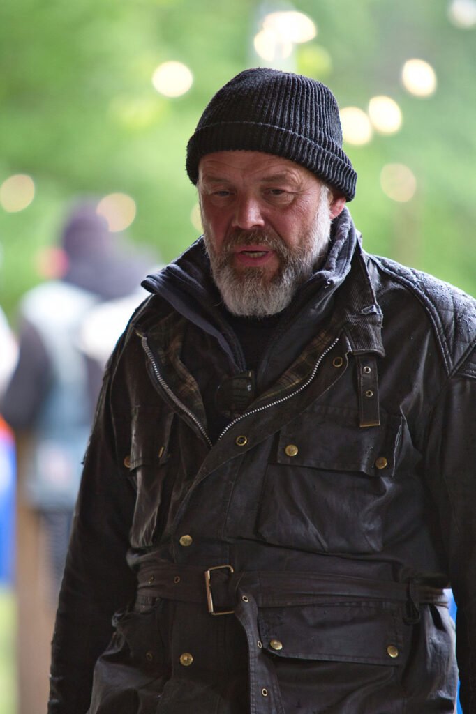 Ein Mann mit grauem Bart, schwarzer Mütze und dunkler Jacke mit Gürtel steht im Freien vor einem unscharfen grünen Hintergrund und Lichtern hinter ihm.