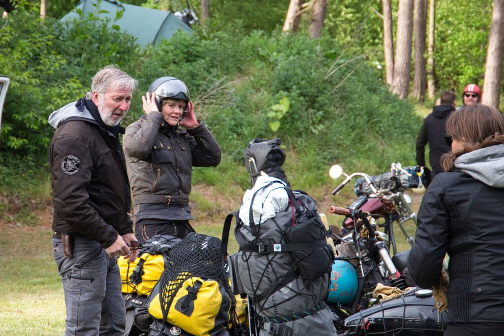 Vier Personen stehen im Freien neben beladenen Motorrädern. Zwei von ihnen tragen Jacken und Helme, einer rückt einen Helm zurecht, und auf den Motorrädern ist Campingausrüstung zu sehen. Der Hintergrund ist grün und bewaldet.
