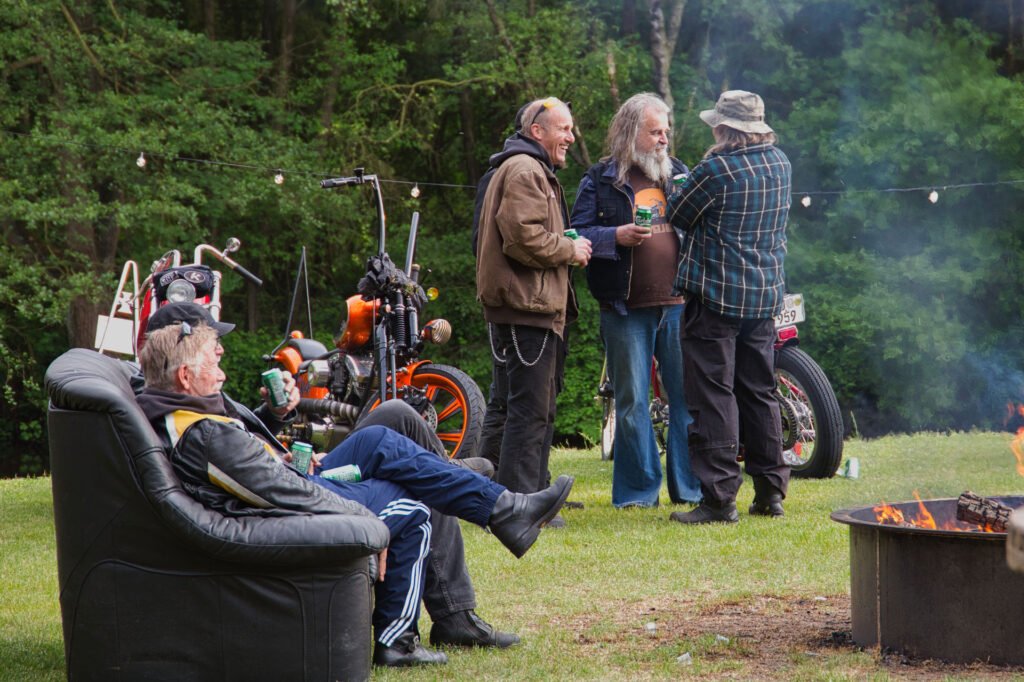 Vier Männer versammeln sich im Freien in der Nähe von Motorrädern und einer Feuerstelle. Drei von ihnen unterhalten sich und halten Getränke in der Hand, während sich einer in einem schwarzen Sessel ausruht. Im Hintergrund sind Bäume und Lichterketten zu sehen.