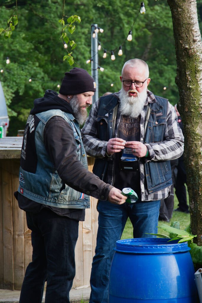Zwei bärtige Männer in Jeanswesten stehen im Freien neben einem blauen Fass, wobei einer eine Getränkedose über das Fass hält. Im Hintergrund sind Lichterketten und grüne Bäume zu sehen.