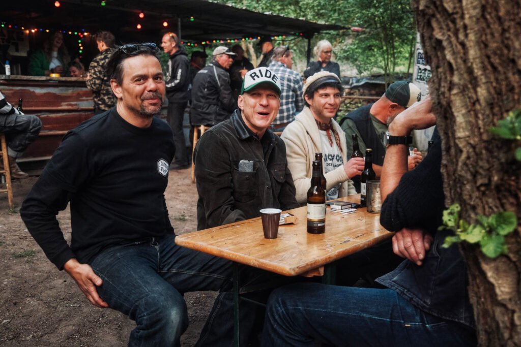 Eine Gruppe von Männern sitzt an einem Holztisch im Freien, lächelt und unterhält sich, auf dem Tisch stehen Getränke und Flaschen. Darüber hängen bunte Lichter, und im Hintergrund sind weitere Menschen in der Nähe eines rustikalen Unterstands versammelt.
