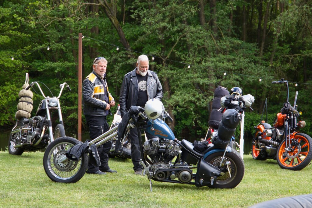 Zwei Männer in Lederjacken stehen draußen im Gras und sehen sich Custom-Motorräder an. Andere Motorräder und Ausrüstungen stehen in der Nähe. Im Hintergrund sind Bäume und Lichterketten zu sehen, die auf ein Treffen im Freien oder eine Motorradveranstaltung hindeuten.