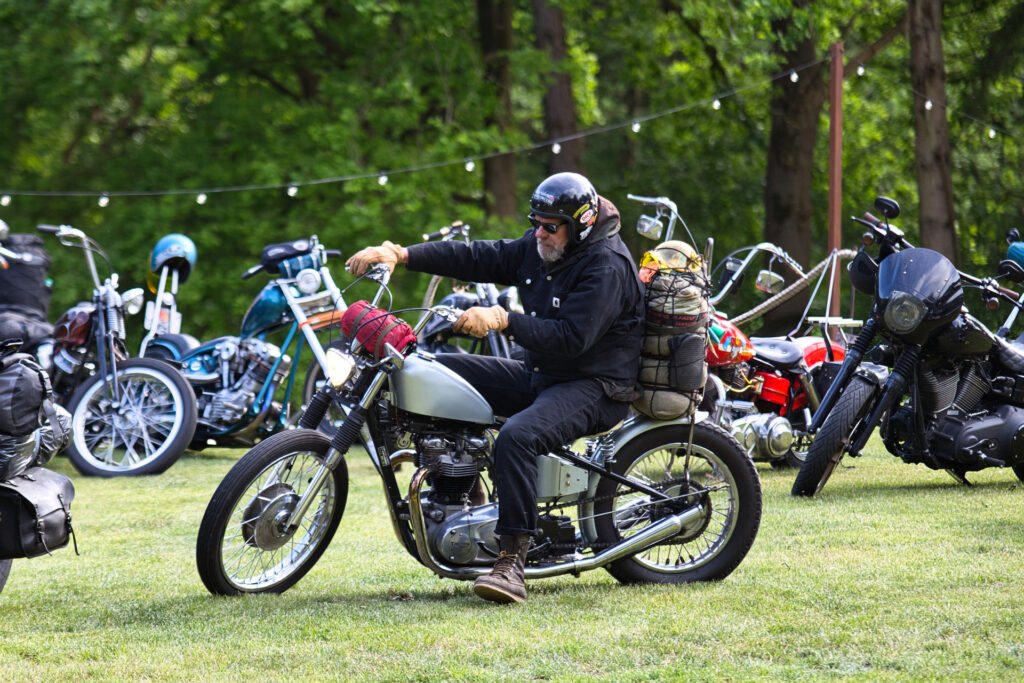 Ein Mann mit schwarzem Helm und schwarzer Jacke fährt auf einem alten Motorrad auf einer Wiese, umgeben von anderen geparkten Motorrädern und Bäumen im Hintergrund. Die Campingausrüstung ist an sein Motorrad geschnallt.