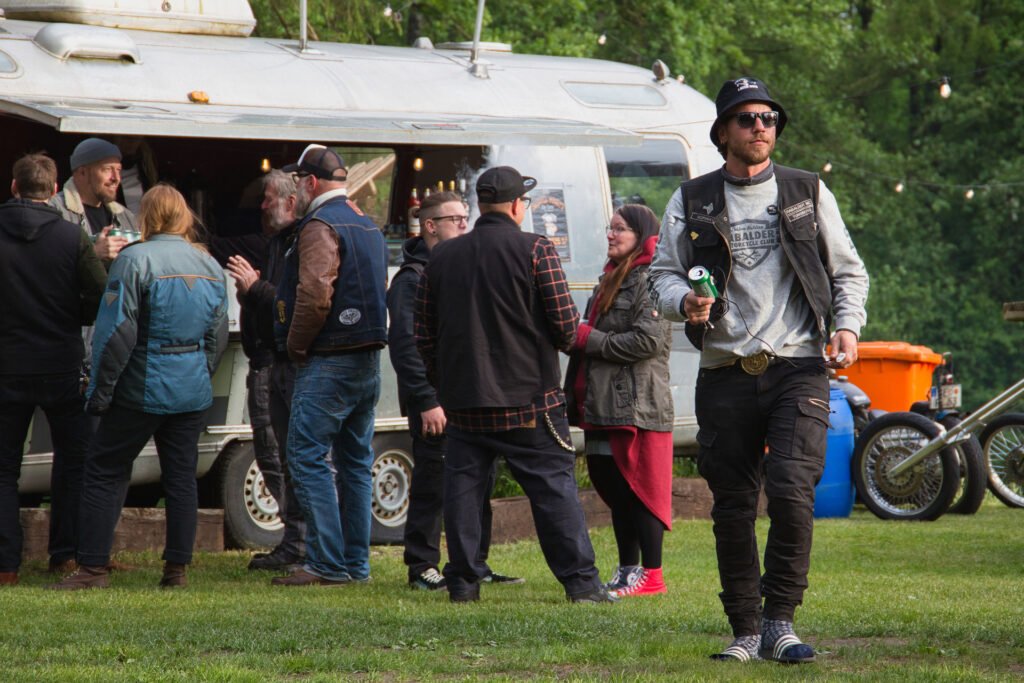 Ein Mann mit Sonnenbrille und Mütze hält ein Getränk in der Hand, während er bei einem Treffen im Freien über das Gras läuft; die Leute unterhalten sich in der Nähe eines Airstream-Anhängers im Hintergrund.