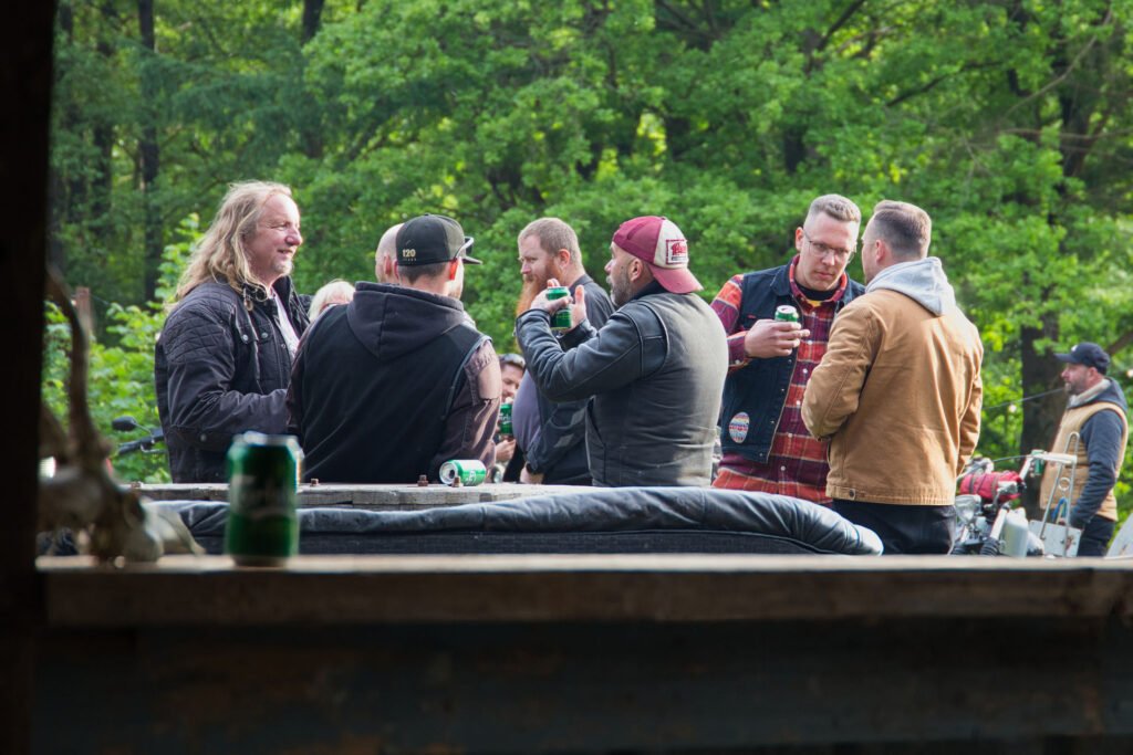 Eine Gruppe von Männern in legeren Jacken steht im Freien, unterhält sich und trinkt aus Dosen, umgeben von Bäumen und Grünzeug. Eine Dose steht auf einer nahe gelegenen Fläche im Vordergrund.