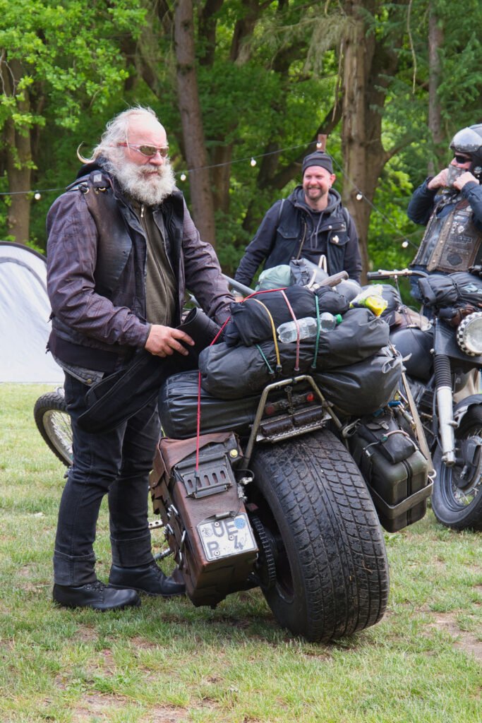 Ein älterer Mann mit weißem Bart steht neben einem schwer beladenen Motorrad, er trägt eine Lederjacke und Stiefel. Im Hintergrund sind zwei weitere Motorradfahrer und Bäume zu sehen. Die Szene scheint bei einer Versammlung im Freien zu spielen.