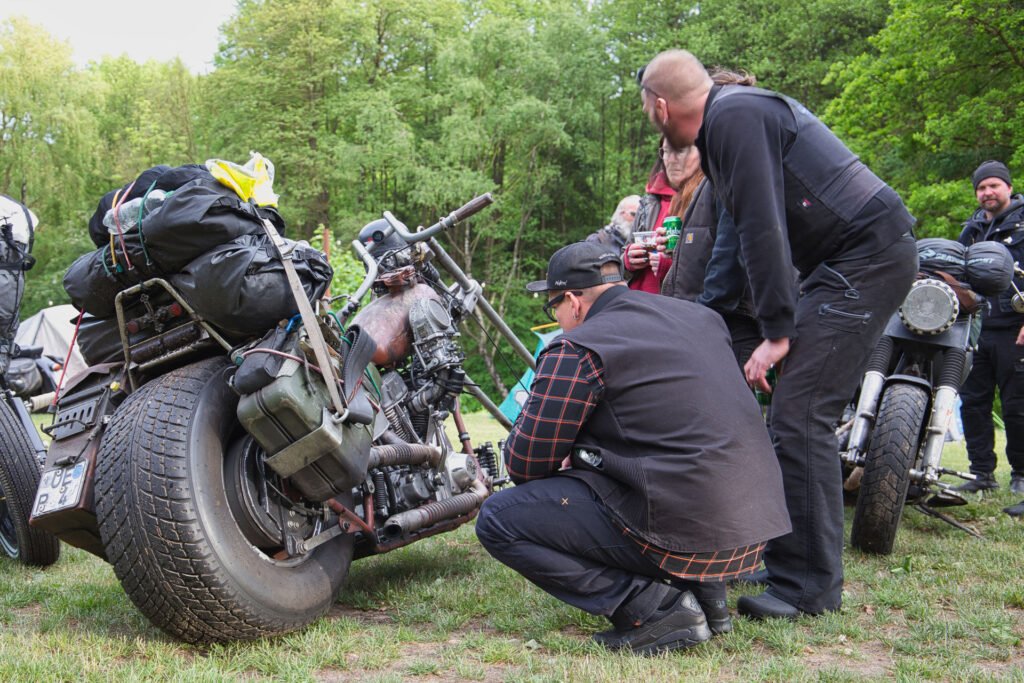 Eine Gruppe von Menschen versammelt sich im Freien um ein altes Motorrad und untersucht dessen Motor. Das Motorrad ist mit Ausrüstung beladen und auf einer Wiese geparkt, mit Bäumen und anderen Motorrädern im Hintergrund.