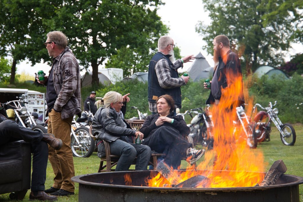 Eine Gruppe von Menschen sitzt und steht um eine große Feuerstelle im Freien, unterhält sich und hält Getränke in der Hand. Im Hintergrund sind Motorräder in der Nähe von Bäumen und Zelten auf einer Wiese geparkt.