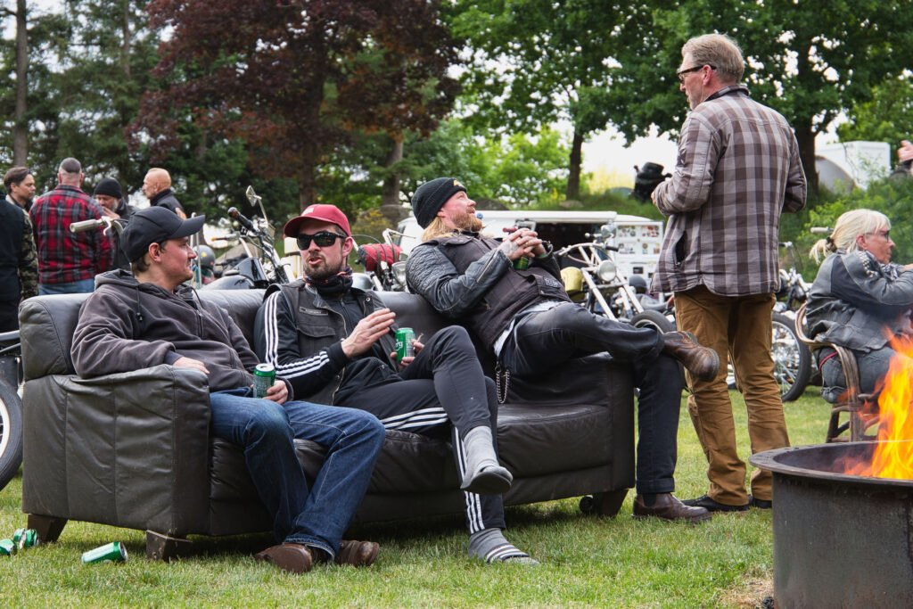 Drei Männer sitzen lässig auf einer Couch im Freien, unterhalten sich und halten Getränke in der Hand. Ein weiterer Mann steht in der Nähe. Im Hintergrund sind Motorräder und weitere Personen zu sehen, und im Vordergrund befindet sich eine Feuerstelle. Es scheint sich um ein geselliges Beisammensein zu handeln.