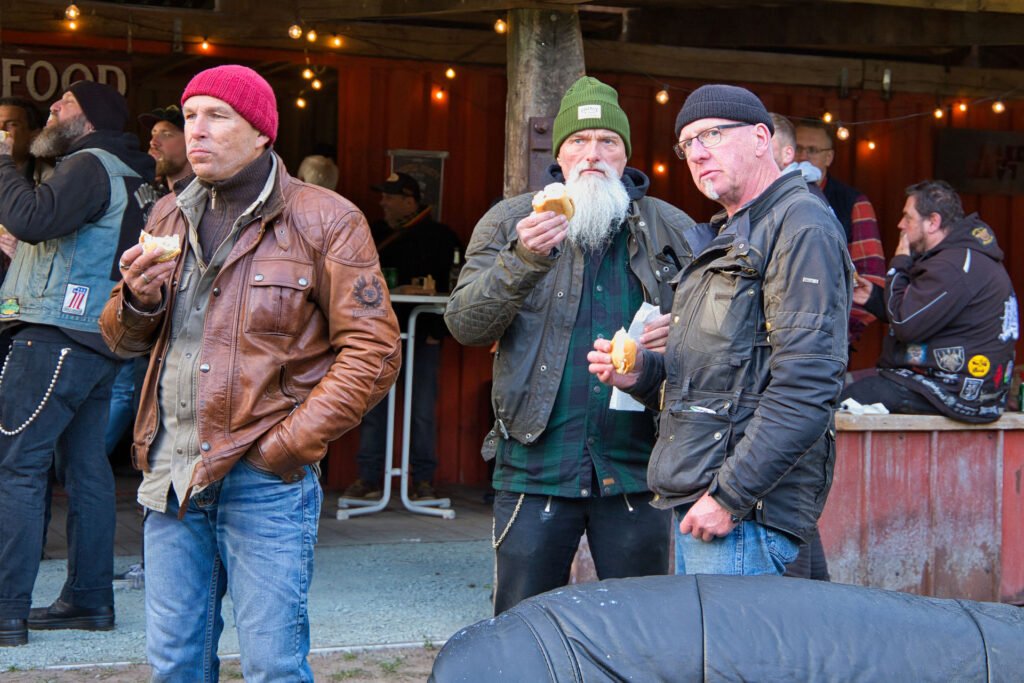 Drei Männer in legeren Jacken und Mützen essen im Freien bei einer rustikalen Veranstaltung, umgeben von anderen Menschen. Im Hintergrund sind Holzstrukturen, Lichterketten und eine entspannte, gesellige Atmosphäre zu sehen.