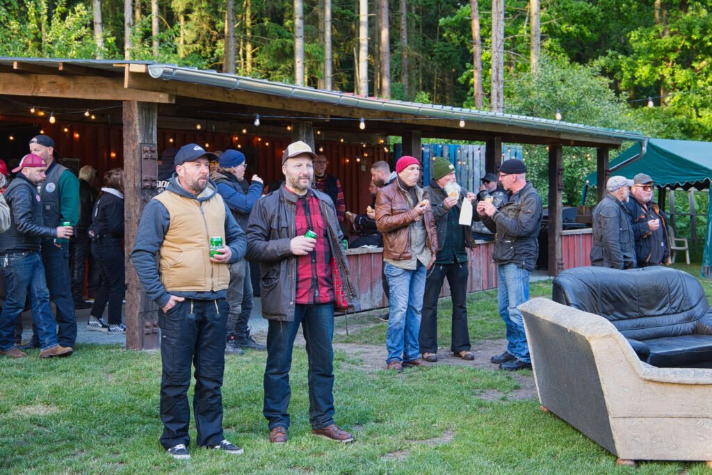 Eine Gruppe von Männern, viele mit Hüten und Jacken, versammelt sich im Freien in der Nähe einer überdachten Holzkonstruktion in einem Waldgebiet. Einige halten Getränke in der Hand, und im Vordergrund steht eine beigefarbene Couch im Gras.