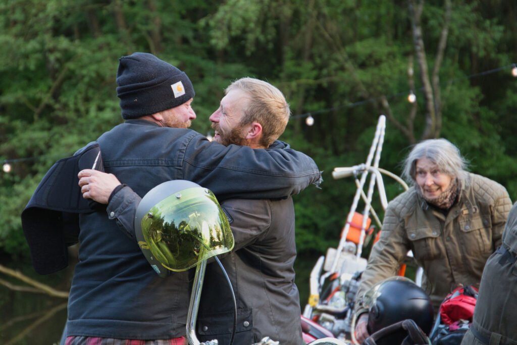 Zwei Männer umarmen sich in der Nähe von Motorrädern in einem Waldstück und lächeln vor Freude. Eine Frau in einer braunen Jacke steht in der Nähe und lächelt ebenfalls. Im Hintergrund hängen Lichterketten zwischen grünen Bäumen.