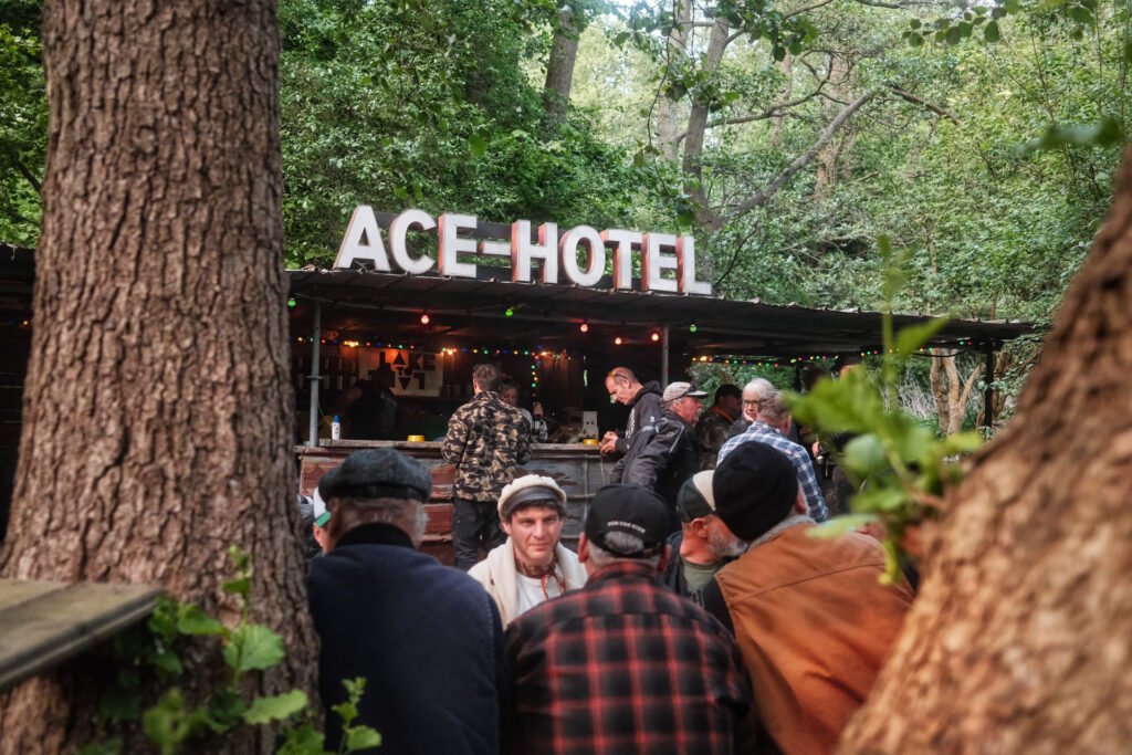 Eine Gruppe von Menschen versammelt sich im Freien an einer rustikalen Bar mit einem großen "ACE HOTEL"-Schild, umgeben von Bäumen und Grünpflanzen, unter einem Baldachin mit Lichterketten.