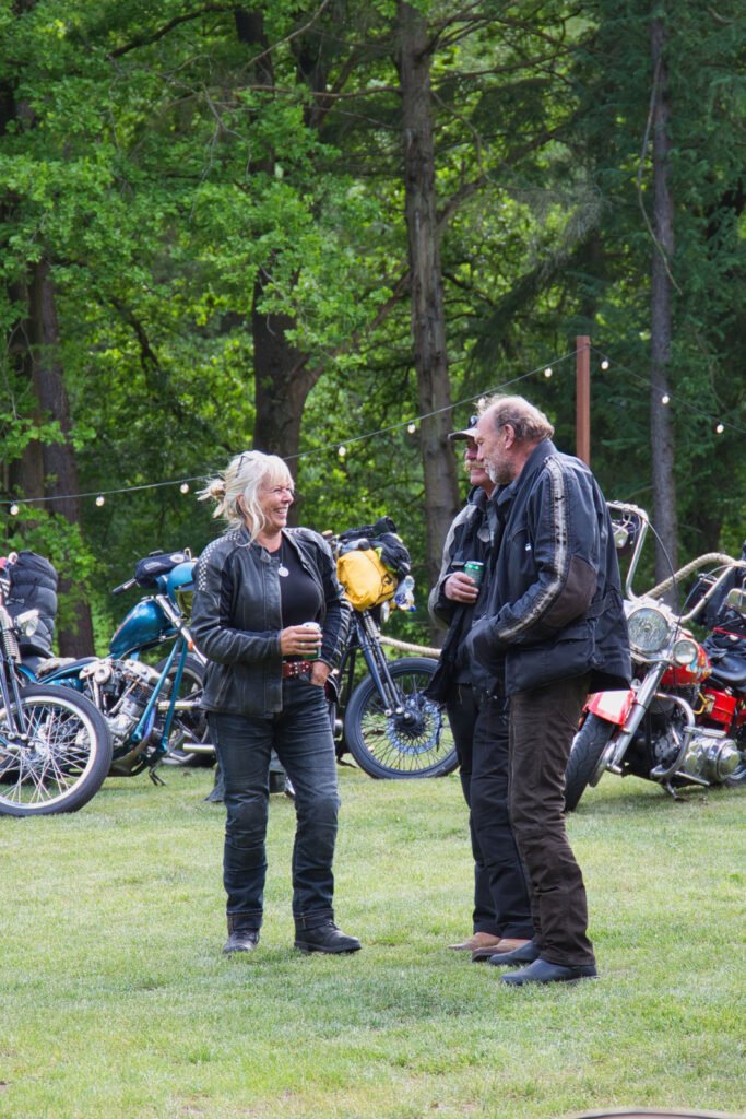 Zwei Personen in Motorradkleidung stehen auf einer Wiese und unterhalten sich mit Getränken in der Hand. Hinter ihnen stehen mehrere geparkte Motorräder und Lichterketten; im Hintergrund sind Bäume zu sehen.