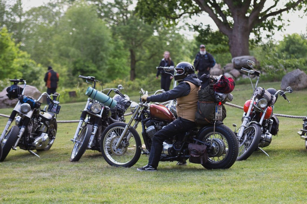 Eine Person mit Helm und Outdoor-Ausrüstung sitzt auf einem verpackten Motorrad inmitten mehrerer geparkter Fahrräder auf einer Wiese, mit Bäumen und Menschen im Hintergrund.