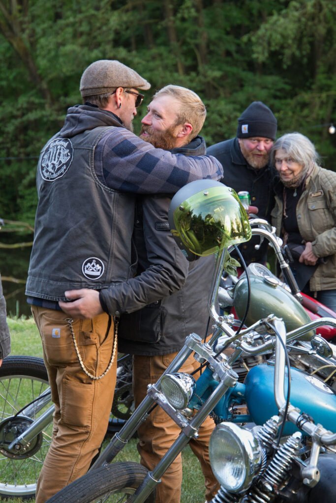 Zwei Männer in lässigen Jacken und Hüten umarmen sich herzlich neben klassischen Motorrädern im Freien, während zwei Personen im Hintergrund lächeln und zusehen. In der Umgebung sind Bäume und Gras zu sehen.