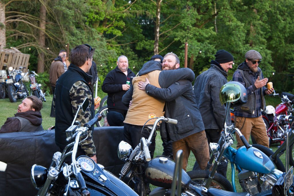 Eine Gruppe von Menschen versammelt sich im Freien zwischen Motorrädern. Zwei Männer in der Mitte umarmen sich, während andere stehen, sich unterhalten und Getränke halten. Der Hintergrund ist mit Bäumen und Grünpflanzen gefüllt.