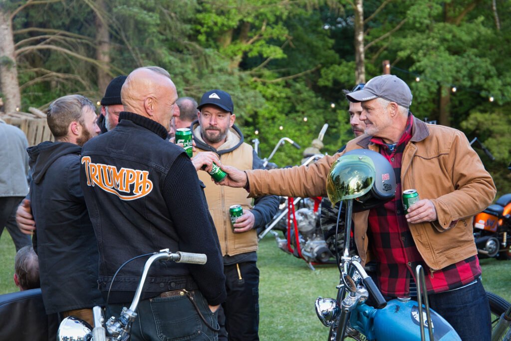 Eine Gruppe von Männern mit Getränken versammelt sich im Freien in der Nähe von Motorrädern. Ein Mann in hellbrauner Jacke und Mütze streckt einem anderen, der eine "Triumph"-Jacke trägt, eine Dose entgegen. Im Hintergrund sind grüne Bäume und mehrere geparkte Motorräder zu sehen.