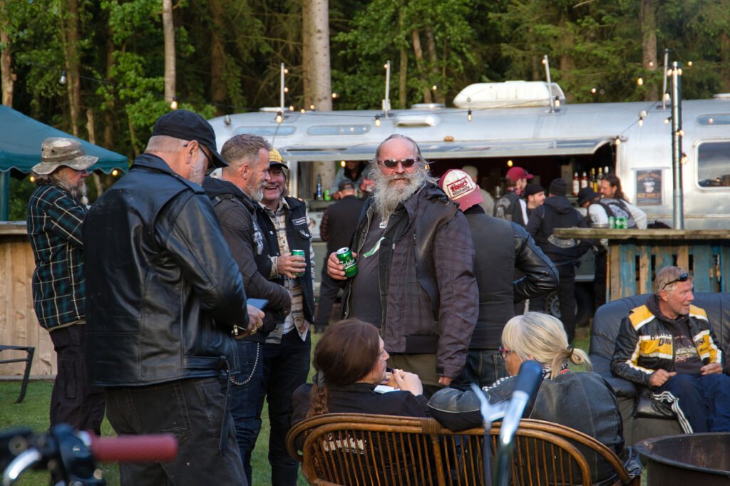 Eine Gruppe von Menschen in Lederjacken trifft sich im Freien in der Nähe eines Wohnwagens, einige stehen, andere sitzen auf Korbstühlen und haben Getränke in der Hand. Es scheint sich um eine zwanglose Zusammenkunft oder ein Fest in einem Waldgebiet zu handeln.