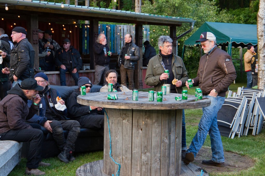 Eine Gruppe von Männern in Outdoor-Jacken versammelt sich um einen großen hölzernen Spulentisch mit Getränken und unterhält sich bei einer Veranstaltung im Freien mit weiteren Personen und einem Zelt im Hintergrund.