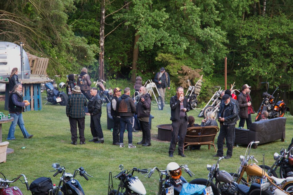 Eine Gruppe von Leuten in Lederjacken versammelt sich im Freien in der Nähe von Motorrädern, plaudert und trinkt. Bäume und Grünflächen umgeben den Bereich, und einige Leute stehen an einer Feuerstelle und an Möbeln im Vintage-Stil.