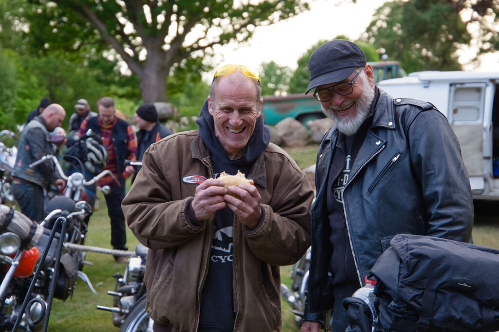 Zwei Männer stehen im Freien neben Motorrädern. Der eine, lächelnd und in einer braunen Jacke, hält ein Sandwich in der Hand. Der andere, mit schwarzer Lederjacke und Mütze, lächelt neben ihm. Im Hintergrund sind Menschen und Bäume zu sehen.