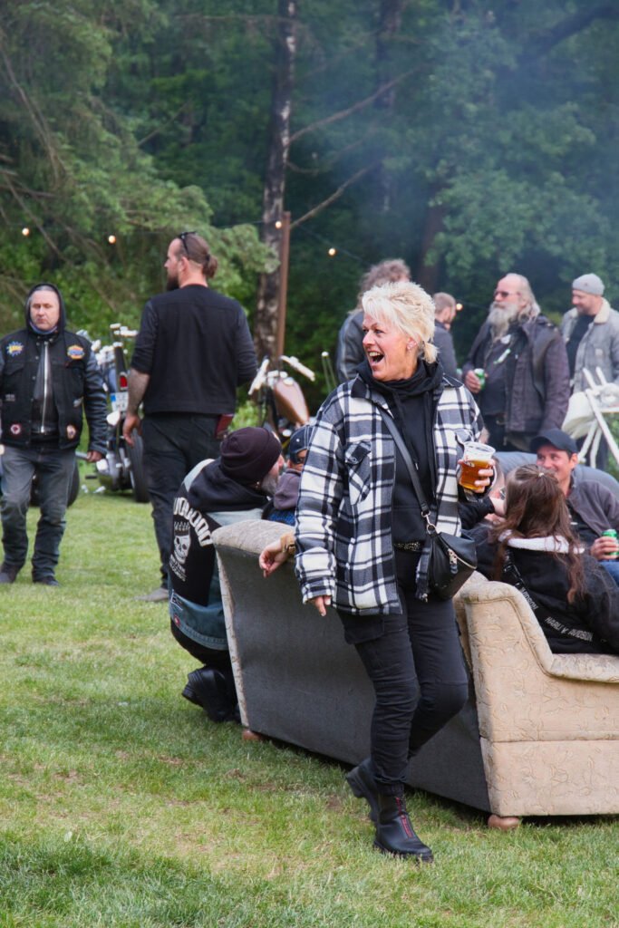 Eine Frau in einer schwarz-weiß karierten Jacke steht lachend mit einem Getränk in der Hand bei einem Treffen im Freien, umgeben von Leuten in Freizeitkleidung und Fahrrädern, von denen einige auf Sofas im Gras sitzen.