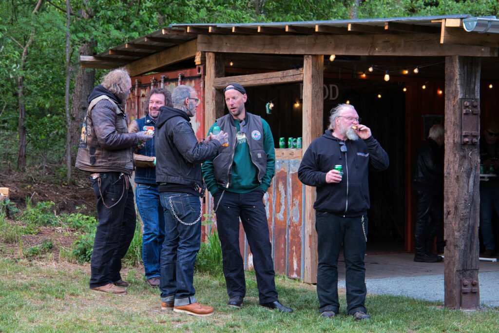 Fünf Männer in legerer Kleidung im Bikerstil stehen vor einer rustikalen Holzhütte, unterhalten sich und trinken Dosengetränke, umgeben von Grün und Bäumen.