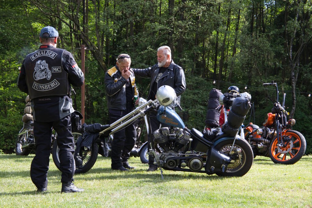Drei Männer in Motorrad-Lederjacken stehen mit ihren Motorrädern im Gras, unterhalten sich und lachen. Im Hintergrund stehen Bäume, und das Sonnenlicht beleuchtet das entspannte Beisammensein im Freien.
