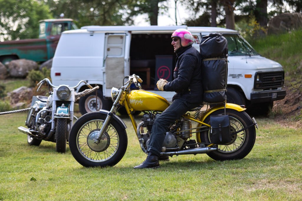 Eine Person mit rosafarbenem Helm und schwarzer Jacke sitzt auf einem gelben Motorrad mit einem großen Rucksack, das auf einer Wiese in der Nähe eines weißen Lieferwagens und eines weiteren Motorrads geparkt ist, wobei im Hintergrund Bäume und Felsen zu sehen sind.