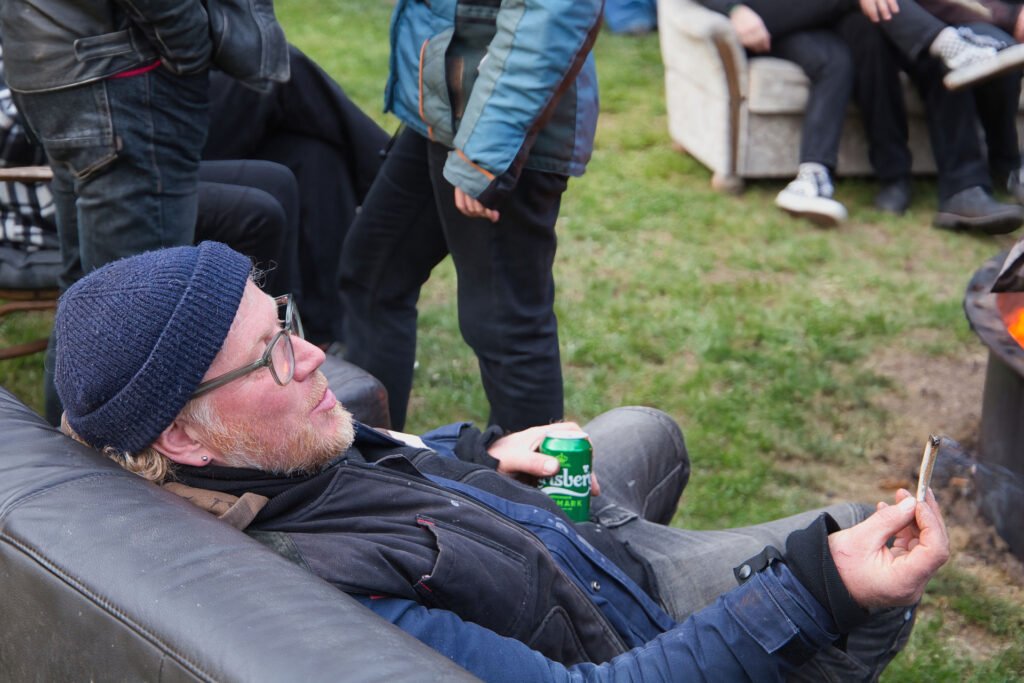 Ein Mann mit Mütze und Brille entspannt sich auf einem Sofa im Freien, in der einen Hand eine Dose Carlsberg-Bier, in der anderen eine Zigarette, während andere in der Nähe im Gras stehen und sitzen.