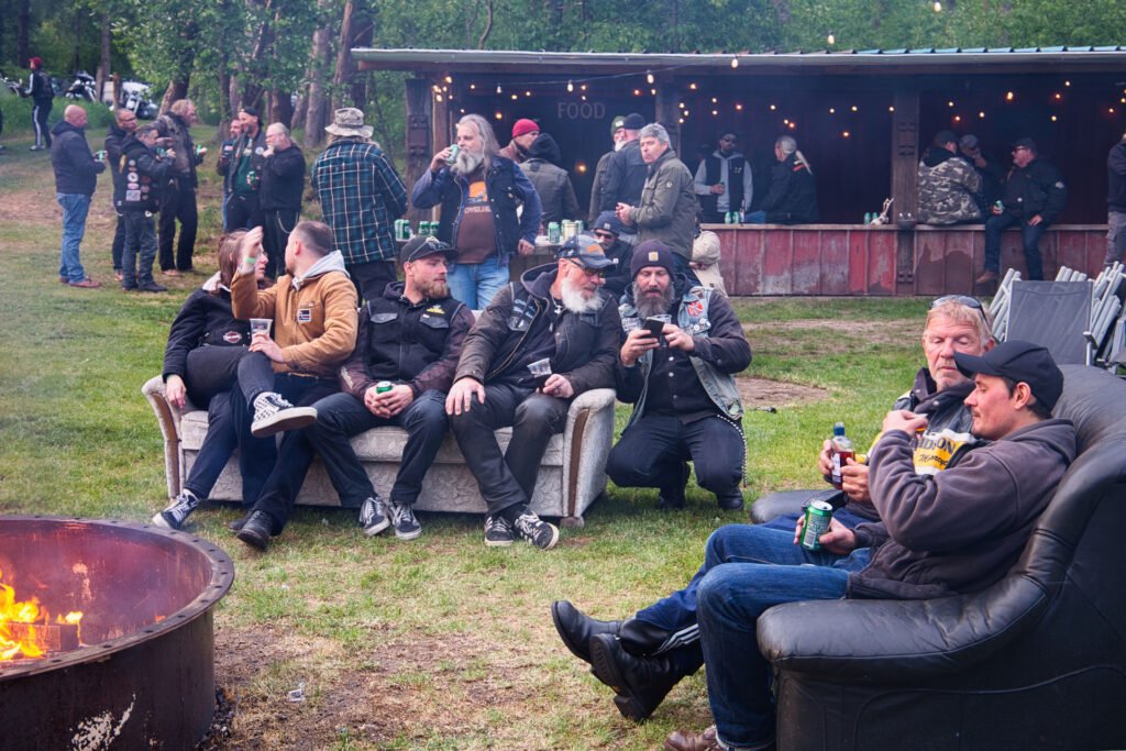Eine Gruppe von Menschen in Jacken und Mützen sitzt auf Sofas in der Nähe einer Feuerstelle bei einem Treffen im Freien, andere stehen und mischen sich im Hintergrund in der Nähe eines Essensstandes unter Lichterketten.
